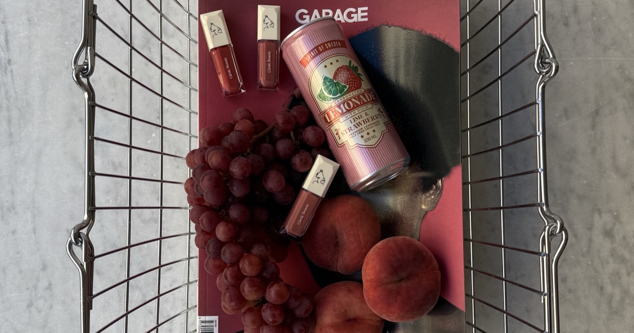 A wire shopping basket holding a magazine, a can of lemonade, lip products, grapes and peaches.