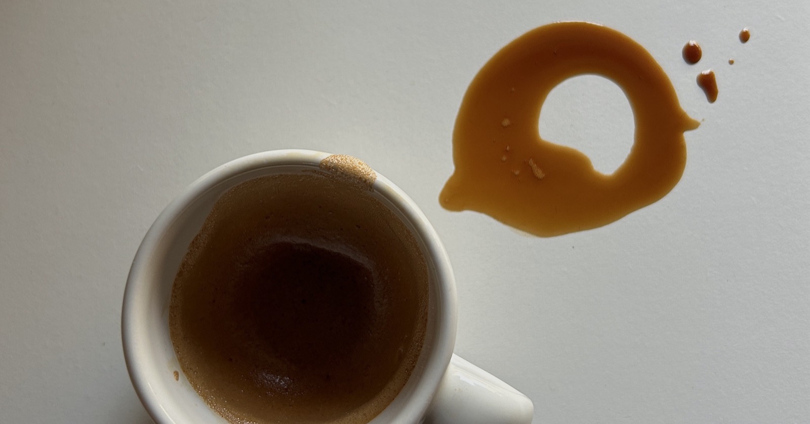 An espresso cup beside a spilt ring of coffee on a white surface.