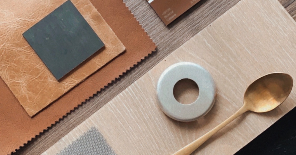 Material swatches laid out on a dark table - leather, linen, paint chips, timber and a brass spoon.