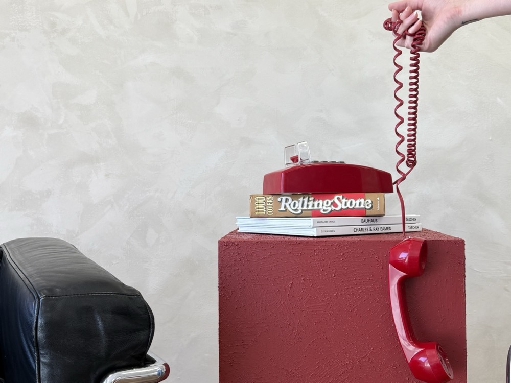 A red rotary phone and design books on a red plinth, handset hanging by its cord.